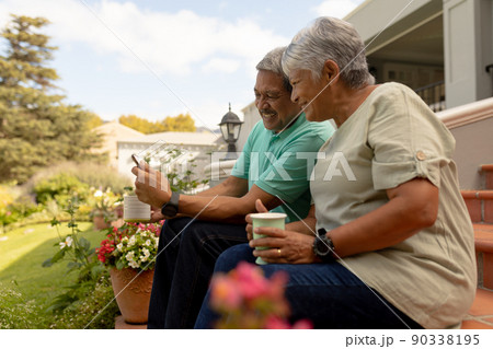 Smiling biracial senior man showing digital tablet to wife holding coffee cup while sitting on steps 90338195