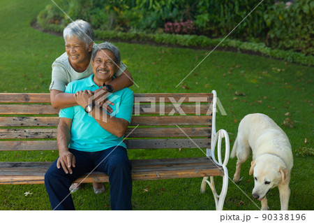 Dog standing by bench and cheerful biracial senior woman embracing husband sitting on bench in park 90338196