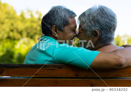 Close-up of smiling biracial senior couple looking at each other while sitting on bench in park 90338202