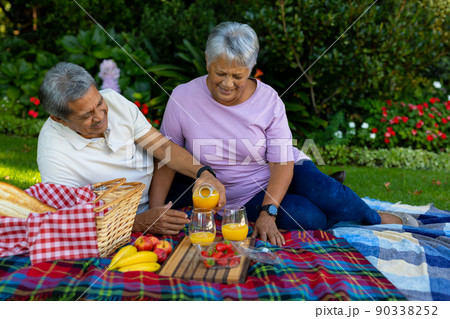 Biracial senior man pouring juice in glass while enjoying picnic with wife against plants in park 90338252