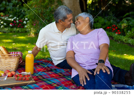 Biracial senior couple looking at each other while sitting with food and drink on blanket in park 90338255