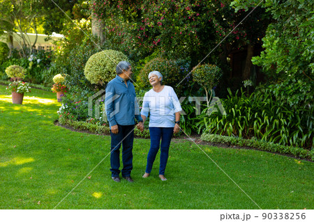 Biracial happy senior woman holding husband's hand while standing against plants and trees in park 90338256