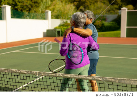 Biracial senior couple with arms around holding rackets and walking at tennis court on sunny day Biracial senior couple with arms around holding rackets and walking at tennis court on sunny day 90338371