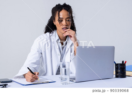 African american mid adult woman writing on clipboard and using laptop against white background 90339068