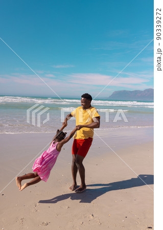 Happy african american young man holding daughter's hands and spinning her at beach against blue sky 90339272