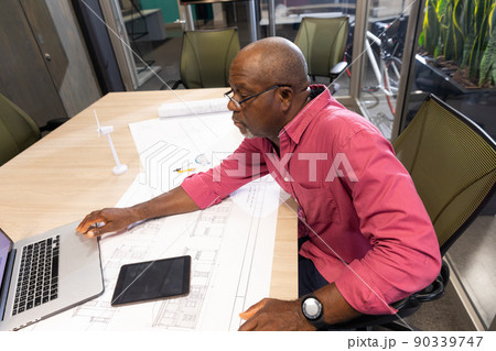 Mature african american engineer using laptop while sitting with blueprint at desk in workplace 90339747