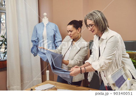 Young Hispanic woman tailor holds fabric and discusses sewing pattern and the quality of textile with a senior fashion designer in a tailoring atelier Young Hispanic woman tailor holds fabric and discusses sewing pattern and the quality of textile with a senior fashion designer in a tailoring atelier 90340051