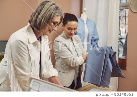 Portrait of a middle-aged Hispanic fashion designer in a stylish jacket holding blue fabric to create a new collection of women's dress shirts in a fashion design workshop 90340053