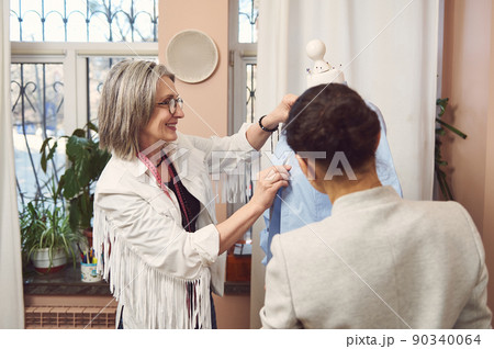 Charming mature female fashion designer and her assistant try on new shirt on mannequin, prepare a new collection of classic clothes for exhibition in fashion design studio Charming mature female fashion designer and her assistant try on new shirt on mannequin, prepare a new collection of classic clothes for exhibition in fashion design studio 90340064