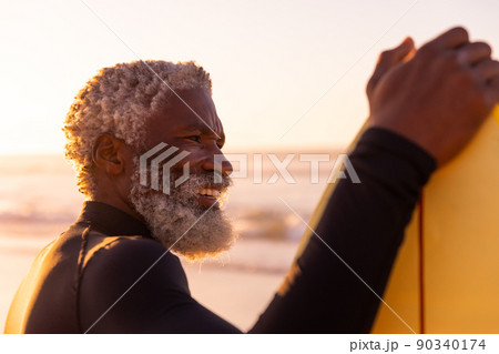 Smiling bearded african american senior man with surfboard looking away at beach against clear sky 90340174