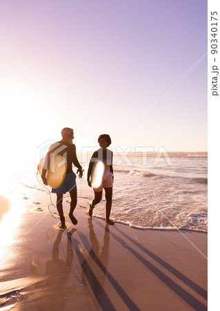 Happy african american couple with surfboards walking at shore against clear sky at sunset 90340175