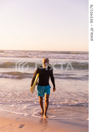 Full length of bearded african american senior man with surfboard walking against sea and clear sky Full length of bearded african american senior man with surfboard walking against sea and clear sky 90340176
