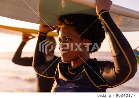 African american mature woman and senior man carrying surfboards on heads at beach at sunset 90340177