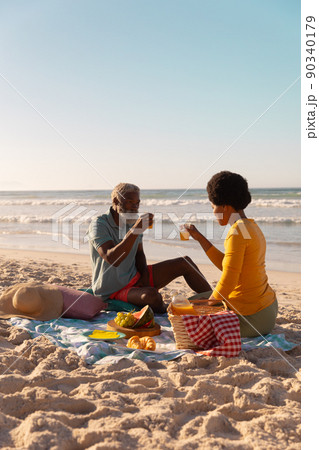 African american couple having juice while sitting with food on blanket at beach against clear sky 90340179
