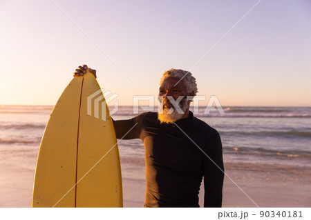 African american bearded senior man with surfboard standing against sea and clear sky at sunset African american bearded senior man with surfboard standing against sea and clear sky at sunset 90340181