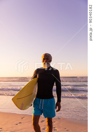 Bearded african american senior man with surfboard standing against sea and clear sky at sunset Bearded african american senior man with surfboard standing against sea and clear sky at sunset 90340182