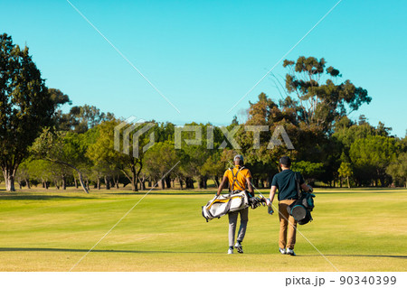 Rear view of multiracial young male friends with golf bags walking at golf course against clear sky Rear view of multiracial young male friends with golf bags walking at golf course against clear sky 90340399