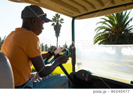 Side view of african american young man wearing cap driving golf cart against sky at golf course 90340477