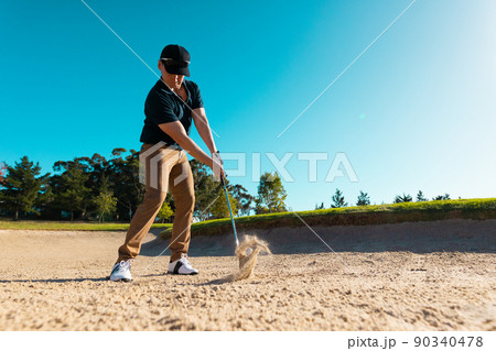 Low angle view of caucasian young man hitting with golf club on sandy land against clear blue sky 90340478