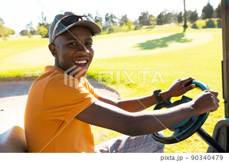 Portrait of happy african american young man wearing cap driving golf cart at golf course Portrait of happy african american young man wearing cap driving golf cart at golf course 90340479