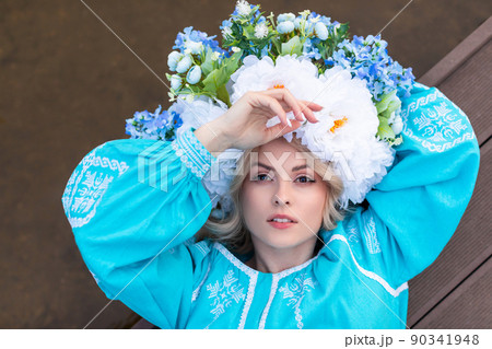 Beautiful young Ukrainian woman dressed in a blue embroidered dress with a large beautiful floral wreath on her head holds a dried flower in her hands and looks at the cameras. The concept of 90341948