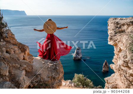 A woman in a flying red dress fluttering in the wind and a straw hat against the backdrop of the sea. 90344208