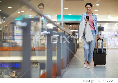Cheerful attractive young lady with coat on arm carrying wheeled luggage and using smartphone while hurrying for boarding onto airplane 90347221