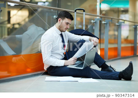 Serious concentrated bearded businessman in formalwear sitting on floor in waiting area of airport and working with papers while using laptop Serious concentrated bearded businessman in formalwear sitting on floor in waiting area of airport and working with papers while using laptop 90347314