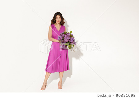 Full-length studio fashion portrait of elegant woman wearing purple dress and holding lilac brunches against white wall. International womans day, Mothers day and spring concept. 90351698