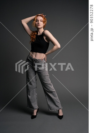 Confident young woman standing in gray studio with hand on waist Confident young woman standing in gray studio with hand on waist 90352278