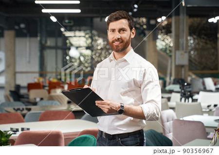 Front view of brunette male with beard standing, holding folder in furniture store. Designer, seller, architect writing, designing, looking at camera. Concept of furniture. Front view of brunette male with beard standing, holding folder in furniture store. Designer, seller, architect writing, designing, looking at camera. Concept of furniture. 90356967