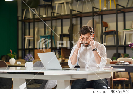 Front view of tired architect, designer sitting at table in furniture shopping. Handsome brunette with beard looking down, holding head by hands, thinking. Concept of shopping. Front view of tired architect, designer sitting at table in furniture shopping. Handsome brunette with beard looking down, holding head by hands, thinking. Concept of shopping. 90356970