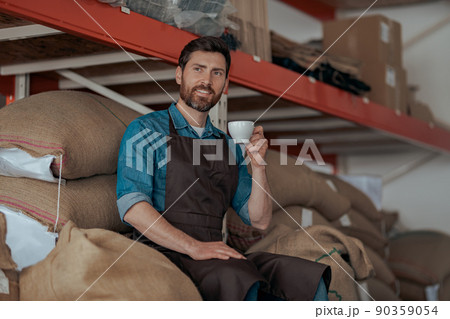 Business owner drinking coffee sitting on warehouse with bags of coffee beans  90359054