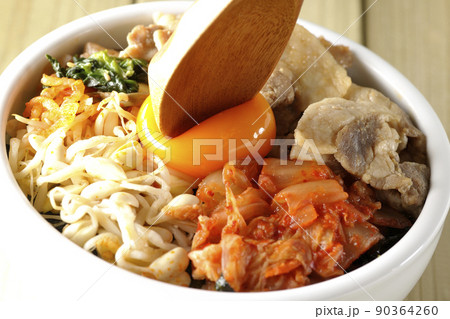 もやしナムルやキムチ、豚肉など沢山の具材が入っているビビンバ丼の卵黄を崩して食べる 90364260