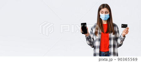 Coronavirus outbreak, working from home, online shopping and contactless payment concept. Unsure girl in medical mask look as making decision at mobile phone, show credit card and smartphone Coronavirus outbreak, working from home, online shopping and contactless payment concept. Unsure girl in medical mask look as making decision at mobile phone, show credit card and smartphone 90365569