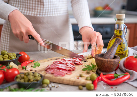Woman slicing Spanish sausage fuet salami with knife on a domestic kitchen Woman slicing Spanish sausage fuet salami with knife on a domestic kitchen 90370656
