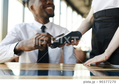 Close-up of smiling black businessman sitting at table and using nfc technology for mobile payment in restaurant Close-up of smiling black businessman sitting at table and using nfc technology for mobile payment in restaurant 90377270