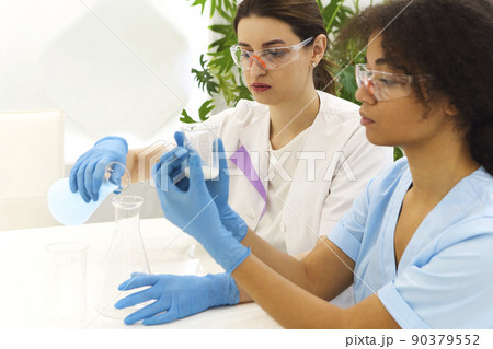Two multi-ethnic female lab scientists in protective uniform carrying out experiments in laboratory 90379552