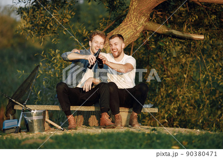 Friends is sitting near a tree and fishing. Two fishermen talking and drinking a beer. One man wearing white t-shirt and other blue shirt. Friends is sitting near a tree and fishing. Two fishermen talking and drinking a beer. One man wearing white t-shirt and other blue shirt. 90380471