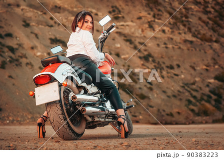 A beautiful adult woman with a high heels and leather pants, sexy posing sitting on motorcycle. Rear view. Rock on the background. The concept of Motorcyclist Day 90383223