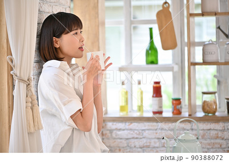 キッチンの壁に寄りかかってマグカップで飲み物を飲む若い女性 90388072