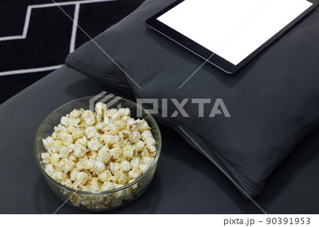 Glass cup with salted popcorn & digital tablet in a gray armchair. Glass cup with salted popcorn & digital tablet in a gray armchair. 90391953