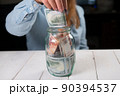 Close-up of a young woman putting hundred-dollar bills in a glass jar. 90394537