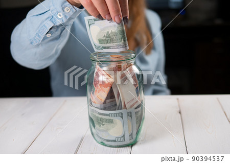 Close-up of a young woman putting hundred-dollar bills in a glass jar. Close-up of a young woman putting hundred-dollar bills in a glass jar. 90394537