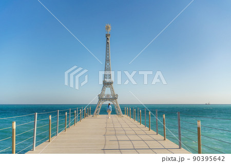 Large model of the Eiffel Tower on the beach. A woman walks along the pier towards the tower, wearing a blue jacket and white jeans. Large model of the Eiffel Tower on the beach. A woman walks along the pier towards the tower, wearing a blue jacket and white jeans. 90395642