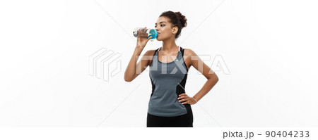 Healthy and Fitness concept - beautiful African American girl in sport clothes drinking water after workout. Isolated on white studio background Healthy and Fitness concept - beautiful African American girl in sport clothes drinking water after workout. Isolated on white studio background 90404233