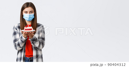 Coronavirus outbreak, lifestyle during social distancing and holidays celebration concept. Cute young woman in medical mask celebrating birthday alone during quarantine, hold b-day cake 90404312