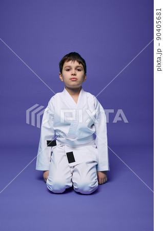 Portrait of a handsome child boy wearing kimono and sitting in aikido stance against purple background with copy space for text. Oriental martial arts concept 90405681