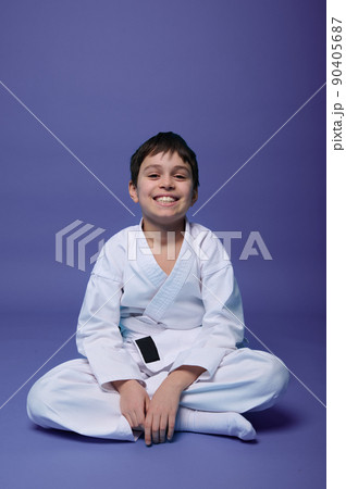 European teenager - aikido fighter - in white kimono sitting in lotus pose while practicing oriental martial arts on purple background with copy space 90405687