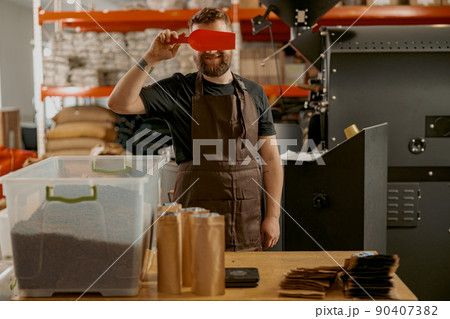 Playful barista closes eyes with a small ladle while standing at a small coffee factory Playful barista closes eyes with a small ladle while standing at a small coffee factory 90407382